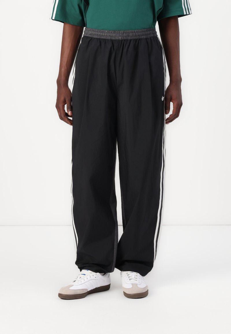 Personne portant un pantalon de survêtement noir avec des bandes blanches sur les côtés, des baskets blanches et un t-shirt vert à manches courtes, debout devant un fond blanc.