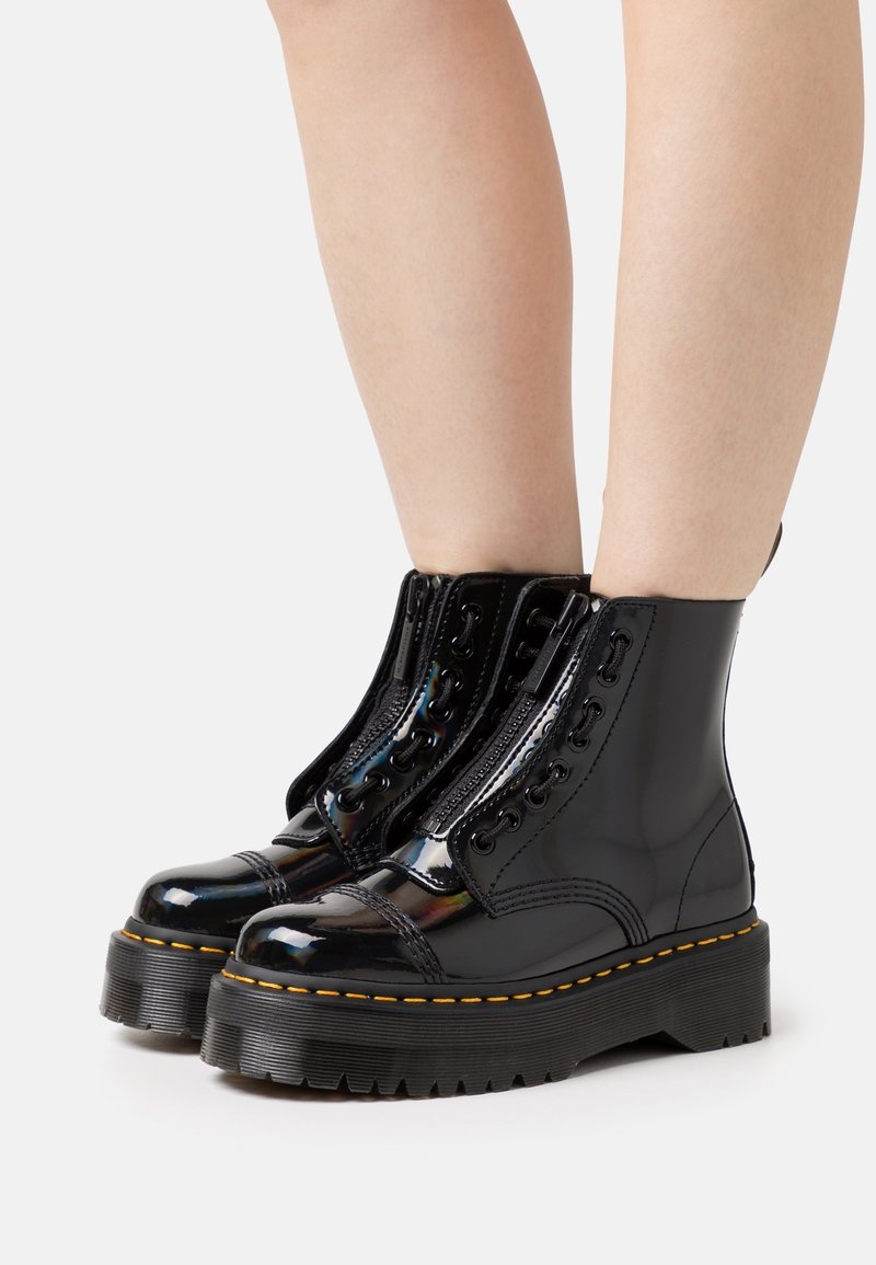Bottines à plateforme en cuir verni noir avec semelles épaisses, fermetures éclair devant et coutures jaunes portées sur jambes nues sur fond blanc.