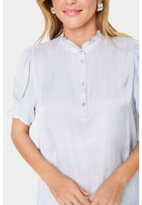 Ljusblå blus i ett slätt tyg med en hög volangkrage, korta puffärmar och knappstängning fram.