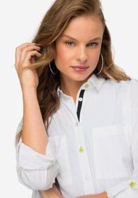 Camicia bianca con bottoni a pressione, caratterizzata da due tasche sul petto, bottoni neri e gialli a contrasto e maniche arrotolate. Tessuto liscio con una texture minimale.