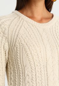 Krémový pleten ý svetr s kabelovými vzory, kulatým výstřihem a žebrovanými detaily. Měkká textura a střední hmotnost.