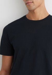 Hombre con una camiseta negra lisa de manga corta y cuello redondo frente a una pared blanca.