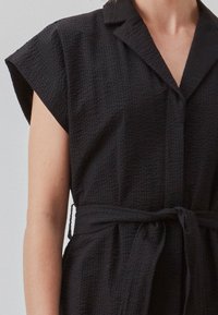 Robe noire texturée avec col, manches courtes et taille nouée. Le tissu présente un motif gaufré et une coupe ample.