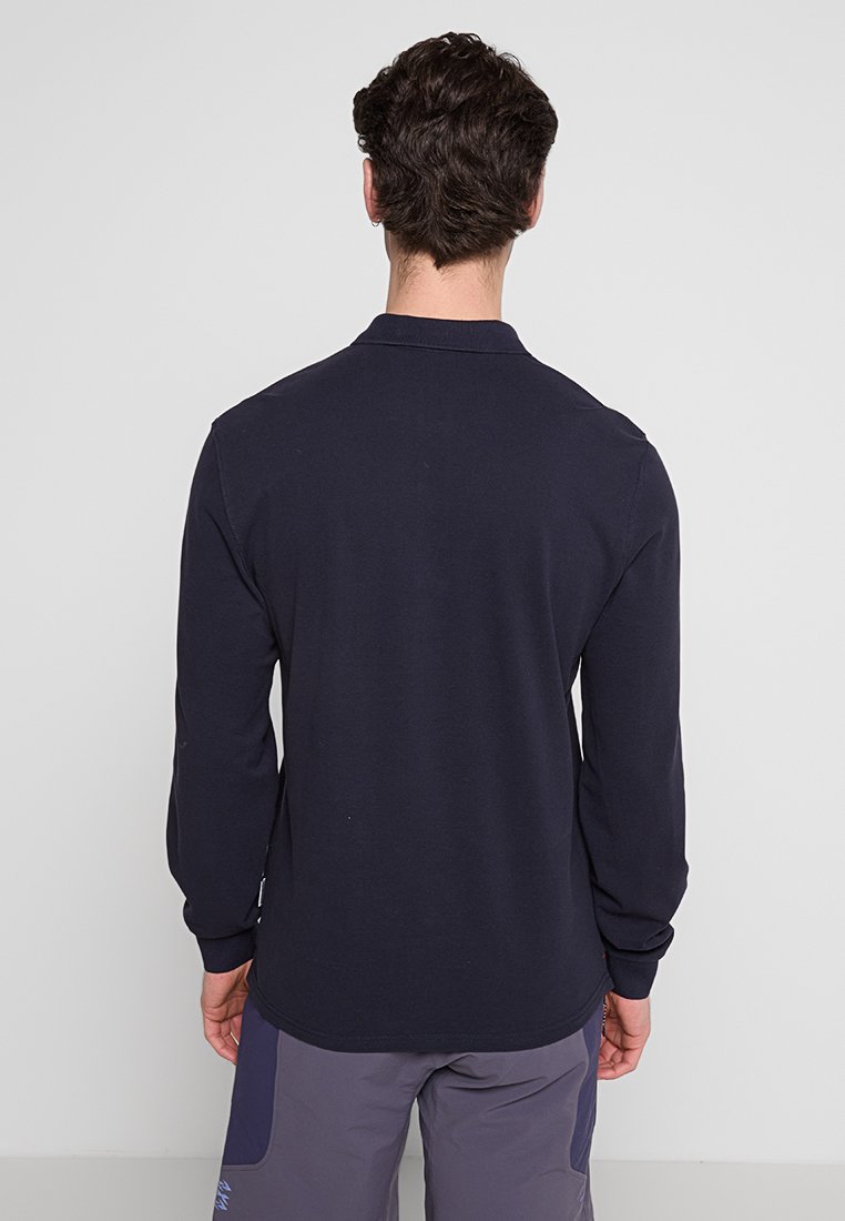 Mężczyzna stojący tyłem do kamery, ubrany w granatową koszulę polo z długim rękawem i szare spodnie, na jednolitym jasnym tle.