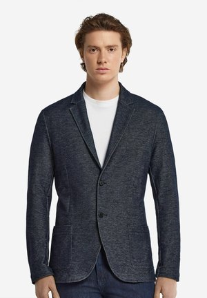 Marineblauer Blazer mit zwei Vordertaschen, Ein-Knopf-Verschluss und einem weichen, strukturierten Stoff. Getragen über einem weißen Hemd.
