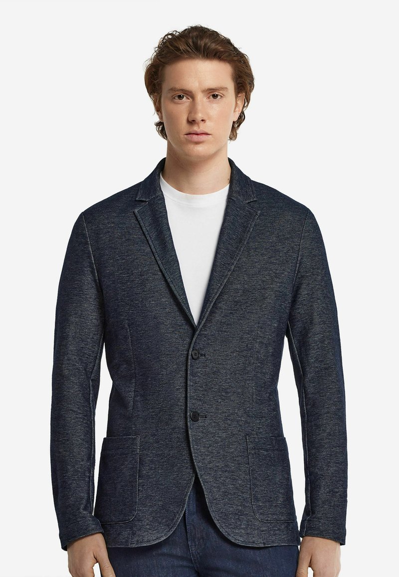 Marinblå kavaj med två framfickor, enfaldig stängning och ett mjukt, texturerat tyg. Bärs över en vit skjorta.