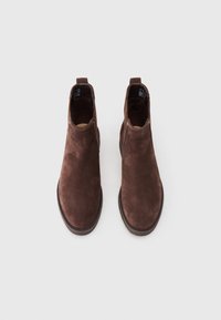 Um par de botas Chelsea de camurça castanha com bicos arredondados e painéis laterais elásticos pretos, vistas de cima sobre um fundo branco.