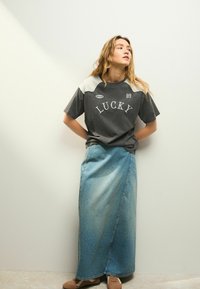 Chemise grise à manches courtes avec le texte "LUCKY", associée à une longue jupe maxi en denim bleu clair. Chaussures brunes à lacets, se tenant devant un fond neutre.