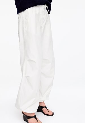 Personne portant un pantalon blanc ample avec taille à cordon et des sandales compensées noires sur fond blanc.
