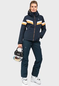 Navy wasserfester Skijacke mit orange und weißen Streifen, kombiniert mit navy Skihosen und weißen Skischuhen, hält einen Helm mit Skibrille.