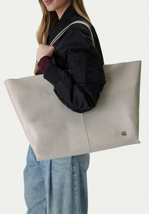 Mujer con chaqueta negra y jeans azules, llevando un gran bolso tote gris sobre el hombro, frente a un fondo liso.