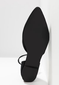 Chaussure en daim noire à bout pointu avec une fine bride à la cheville et une semelle extérieure en caoutchouc texturé, présentant un design minimaliste et une étiquette de taille 37.