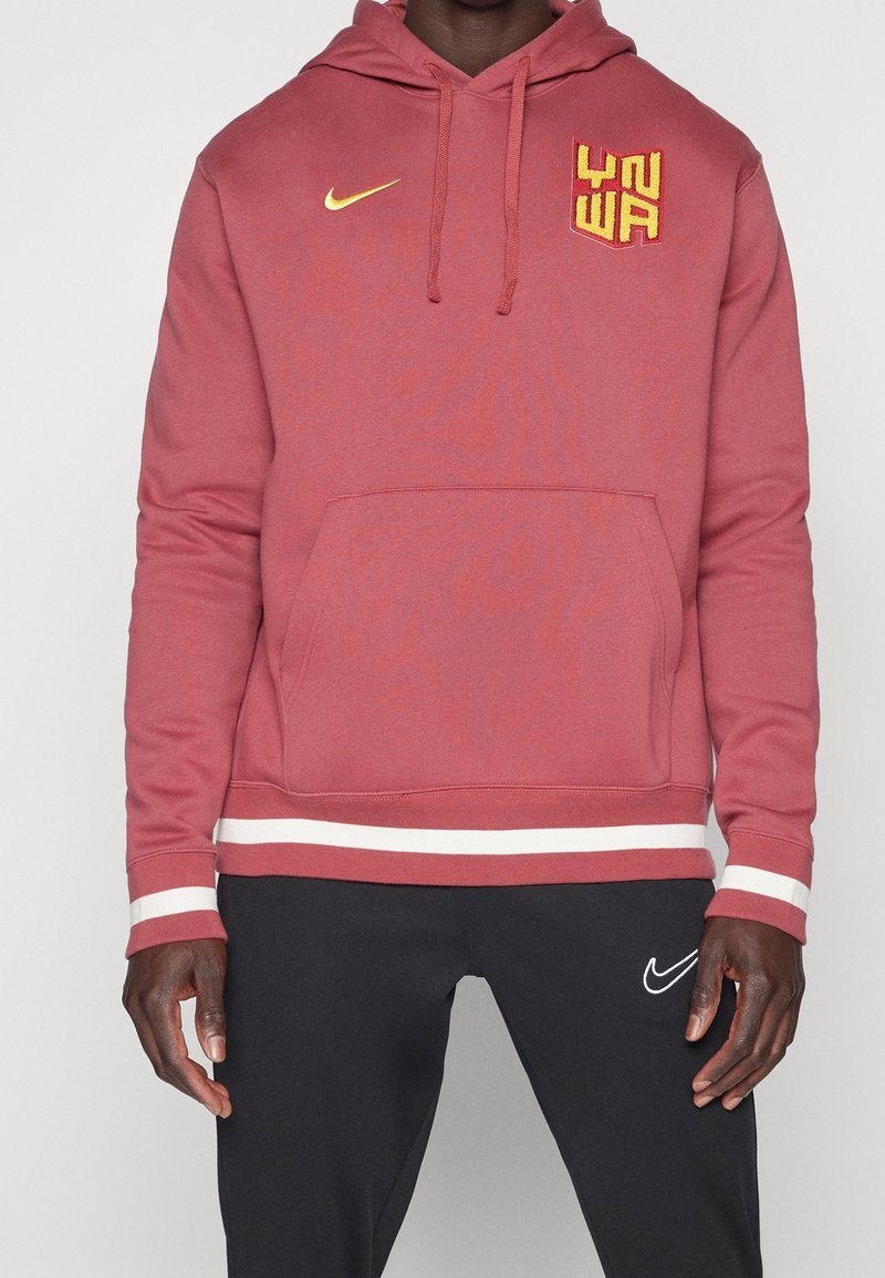 Homme portant un sweat à capuche rouge rouille avec poche frontale et logo jaune, assorti à un pantalon de sport noir Nike avec logo swoosh blanc.