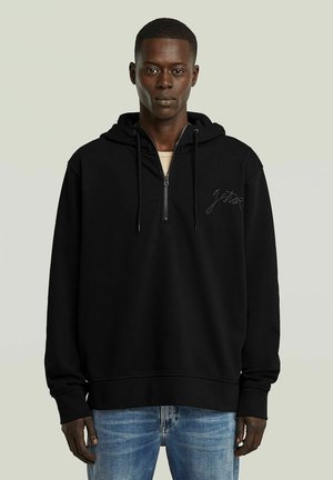 Junger Mann trägt einen schwarzen Kapuzenpullover mit halbem Reißverschluss und Kordeln sowie hellblaue Jeans, steht vor einem schlichten, hellen Hintergrund.