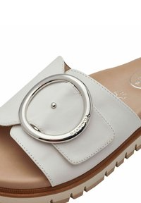 White leather slide sandal featuring a large metallic circular accent, textured upper with stitching detail, and a chunky, textured sole.