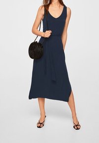 Navy blue sleeveless dress with a V-neck, waist tie, side slit, and a smooth texture, paired with a round black bag and strappy sandals.