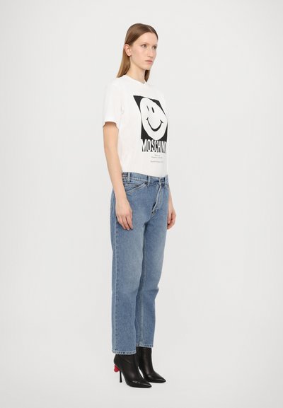 Balta grafika marškinėliai su juodu šypsenos veidu ir užrašu "MOSCHINO", derinami su šviesiai mėlynomis tiesiomis kelnėmis ir juodais aukštakulniais batais.