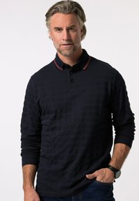 Camisa polo de manga larga color marino con un patrón texturizado, con tapeta de dos botones y acentos en el cuello a rayas rojas y blancas. Se lleva con jeans oscuros.