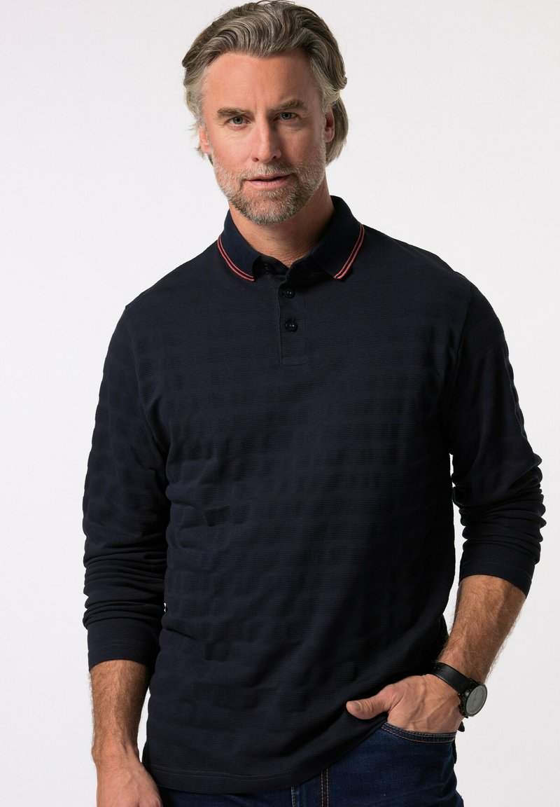 Camisa polo de manga larga color marino con un patrón texturizado, con tapeta de dos botones y acentos en el cuello a rayas rojas y blancas. Se lleva con jeans oscuros.