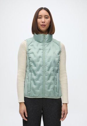 Svetlozelená prešívaná vesta s vysokým golierom, zipsom a bočnými vreckami, nosená cez krémový rebrovaný sveter.