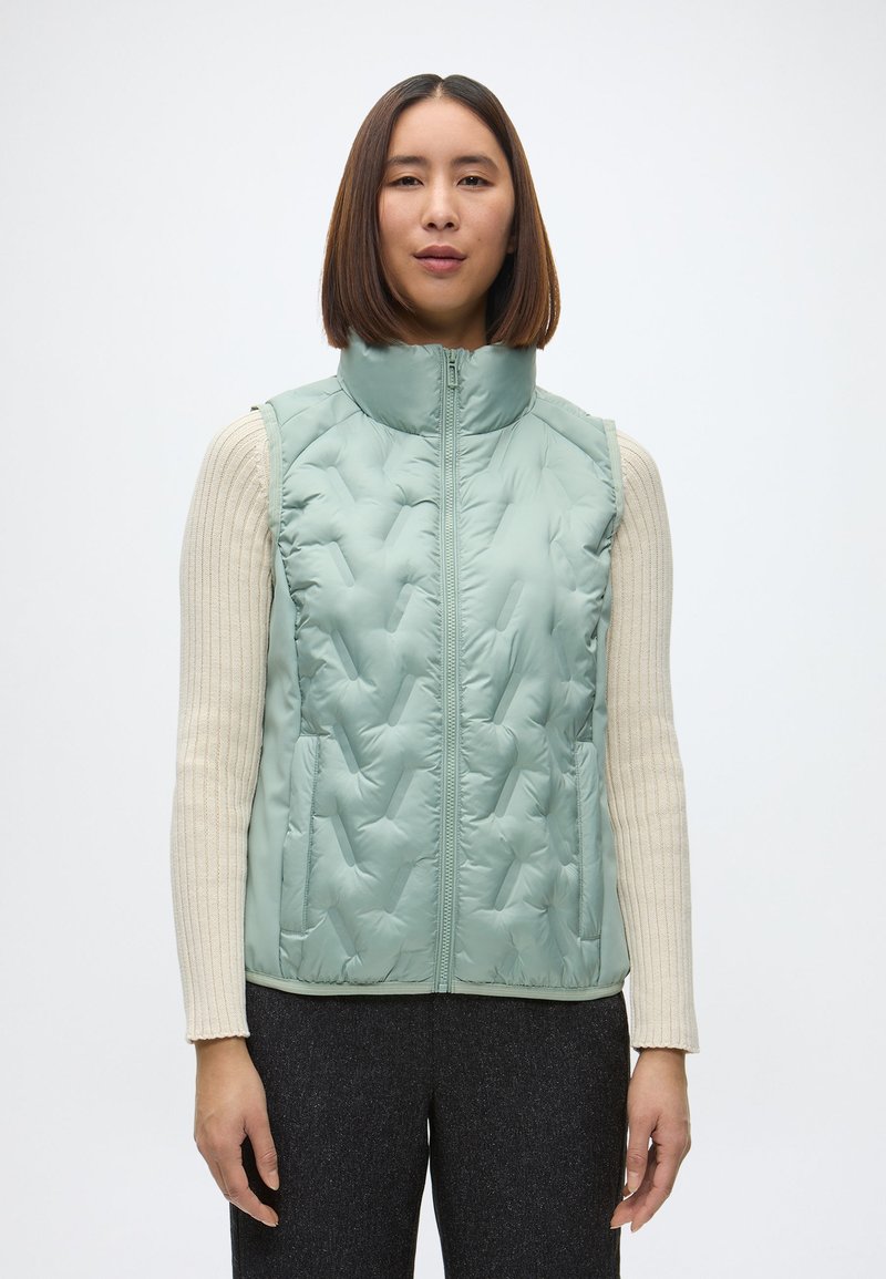 Svetlozelená prešívaná vesta s vysokým golierom, zipsom a bočnými vreckami, nosená cez krémový rebrovaný sveter.