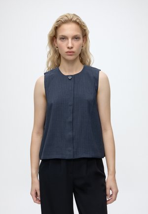 Femme blonde portant un haut bleu marine sans manches à fines rayures avec un seul bouton au col et un pantalon noir, debout sur un fond blanc.