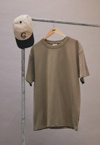 Un tricou din bumbac de culoare verde măslin, cu guler rotund, mâneci scurte și cusături vizibile, atârnă pe un suport metalic; o șapcă de baseball bej atârnă deasupra.