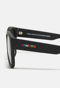 Occhiali da sole neri realizzati in plastica riciclata, con finitura opaca e logo "ARNETTE" colorato sul lato. Dettaglio a colori sfumati sul logo.