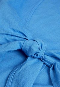 Top de tela azul con un detalle anudado en la cintura, con una textura suave y una apariencia ligera y transpirable.