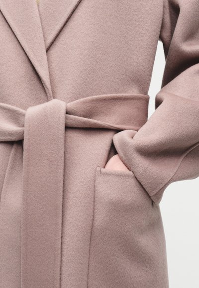 Μαλακό ροζ μάλλινο παλτό με ζώνη δεμένη και χέρι που έχει μπει στην μπροστινή τσέπη σε απλό φωτεινό φόντο.
