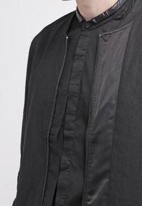 Giacca bomber nera con una texture liscia, colletto stand-up, chiusura frontale con zip e dettagli in tessuto a contrasto sulle spalle.