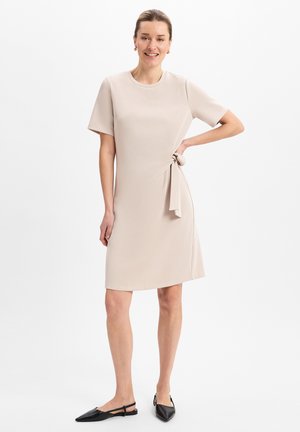 Beiges Kurzarmkleid mit rundem Ausschnitt und seitlicher Knotendetail. Glatter Stoff, knielang, kombiniert mit schwarzen spitzen Ballerinas.