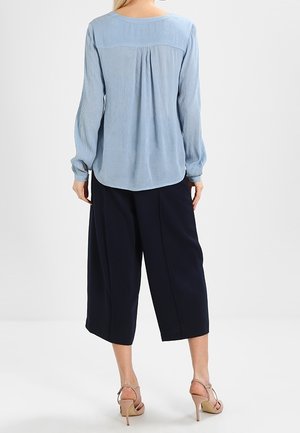 Femme portant un chemisier bleu clair à manches longues, un pantalon noir large et court, et des sandales beiges à talons hauts, debout dos à la caméra.