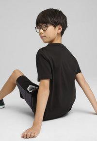 Camiseta negra y pantalones cortos grises con un detalle de bolsillo. El chico lleva gafas, está sentado de lado, con el cabello corto y negro, frente a un fondo neutro.