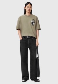 Oversized t-shirt i olivengrøn med kraniegrafik, kombineret med løse sorte jeans og sorte sandaler. Personen holder en sort taske.