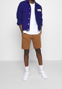 Veste violette à manches longues avec deux poches poitrine sur un t-shirt blanc, associée à un short marron et des baskets blanches, sur fond neutre.