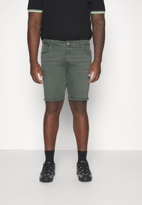 Green denim shorts with a slim fit, rolled hem, and five pockets. Paired with black sneakers and a black short-sleeve shirt.