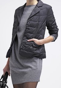 Kvinna i grå stickad klänning och svart vadderad blazer, som håller i en svart handväska med handen i blazerns ficka.