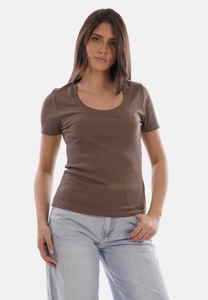 Donna con capelli lisci castani, indossa una maglietta marrone a maniche corte con scollo a barca e jeans azzurri, in piedi con una mano in tasca.