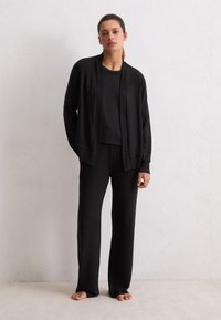 Donna in piedi a piedi nudi contro una parete chiara, indossa un cardigan nero largo, una maglietta nera e pantaloni neri a gamba larga.