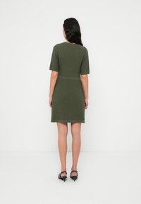 Πλεκτό φόρεμα σε ελιά πράσινο με κοντά μανίκια και εφαρμοστή μέση, με υφασμένο σχέδιο και μήκος μέχρι το μέσο του μηρού. Μαύρα παπούτσια με τακούνι.