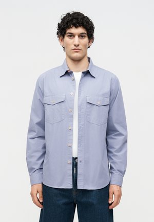 Νέος άνδρας με σγουρά μαλλιά που φοράει ανοιχτό μπλε πουκάμισο πάνω από λευκό μπλουζάκι και σκούρο μπλε τζιν, στέκεται μπροστά σε ένα απλό φόντο.