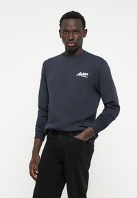 Donkerblauwe sweatshirt van zachte stof met lange mouwen en geribde manchetten, voorzien van een witte geborduurde 'Aspen'-logo op de borst, gecombineerd met zwarte broek.