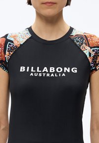 Άνω μέρος του κορμού ενός ατόμου που φοράει μαύρο πουκάμισο Billabong Australia με πολύχρωμα μανίκια σε λουλουδάτο μοτίβο.