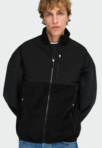 Giovane uomo con capelli biondi scompigliati che indossa una giacca in pile nera con zip e jeans blu, in piedi contro uno sfondo chiaro.