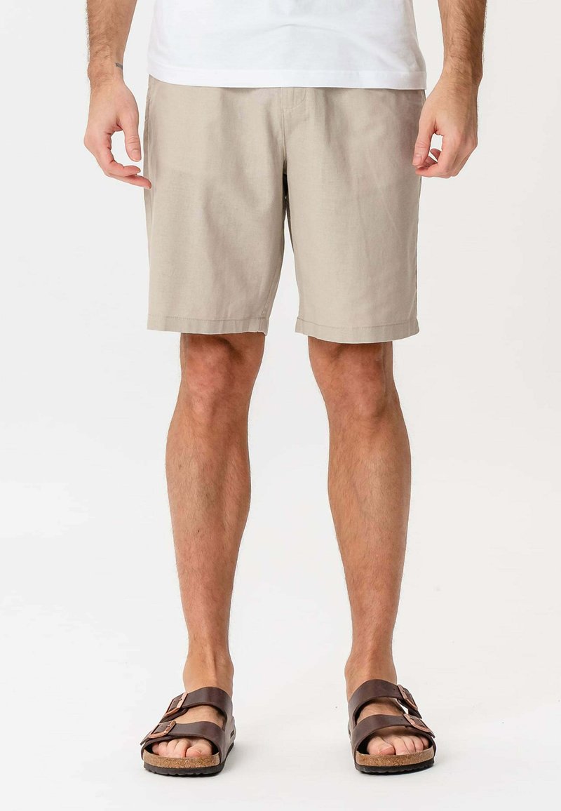 Mand står iført beige shorts, hvid skjorte og brune sandaler med åben tå og to spænder mod en ensfarvet baggrund.