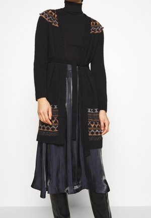 Mujer con un cárdigan de punto negro con hombros y bolsillos estampados, sobre un cuello alto negro y una falda midi negra brillante.