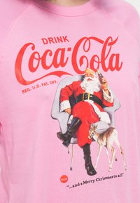 Santa Claus v červenom obleku, ktorý drží fľašu Coca-Coly, sediaci na stoličke, s malým jeleňom vedľa seba a logom Coca-Coly nad ním.