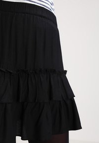 Jupe noire à volants en étages portée avec un haut rayé blanc et bleu marine rentré, montrée de la taille jusqu'au milieu de la cuisse sur fond uni.