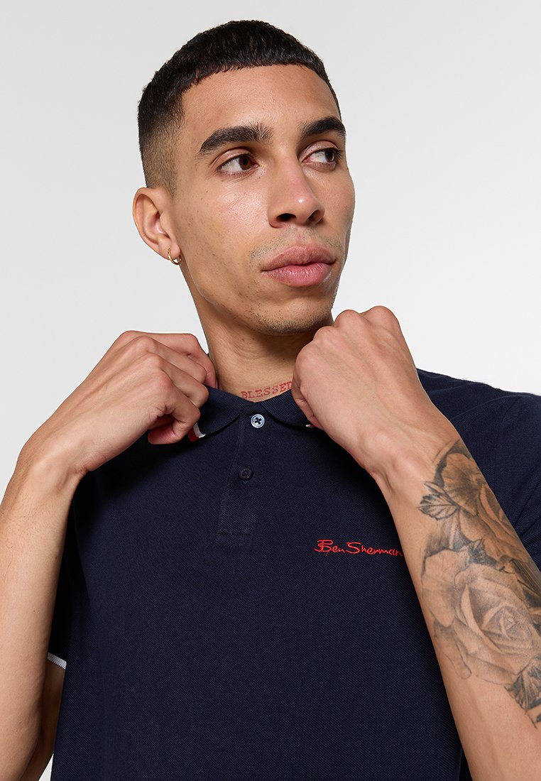 Polo shirt bleu marine avec une finition texturée, patte de boutonnage à deux boutons et logo rouge brodé. Agrémenté de poignets blancs, porté autour du col.